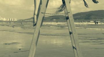 Uma escada está aberta na areia da praia. Uma mulher está com seu dorso flexionado sobre o topo da escada, de modo que seus braços estão de um lado da escada e suas pernas de outro. #Paratodosverem
