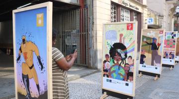 paineis expostos em calçadão. Atrás de um deles aparece a lateral do corpo de uma mulher que fotografa uma das obras. #paratodosverem 