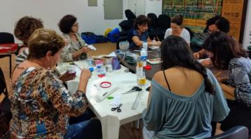 Mulheres estão sentadas em torno de uma mesa fazendo trabalhos manuais. #Pracegover