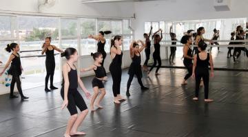 meninas estão em salão em aula de balé. Elas se movimentam em pé em diversas formas. #paratodosverem