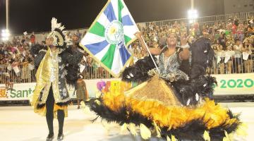 Mestre-sala e porta-bandeira se apresentam em sambódromo.Público está ao fundo. #paratodosverem