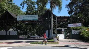 Fachada do jardim botânico com placa, palmeira à frente e árvores variadas ao fundo, atrás do portal de acesso. #paratodosverem