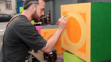 artista de rua está pintando um cubo com desenho da mureta tradicional de santos. #paratodosverem