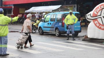 mulher atravessa rua na faixa. Ela puxa um carrinho de feira. Um agente da CET está de um lado da rua fazendo o gesto com o braço esticado. Outro está parado dooutro lado da rua. #paratodosverem