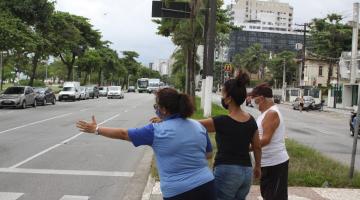 agente da CET faz sinal da faixa viva com o braço estendido e orienta dois pedestres em canteiro central na avenida. #paratodosverem