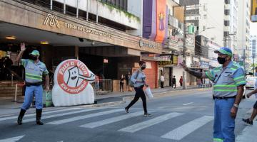 Agentes da CET fazem gesto de mão estendida sobre faixa de pedestre. Ao fundo, uma mulher atravessa a rua. Do lado esquerdo, o inflável da campanha Faixa Viva. #paratodosverem