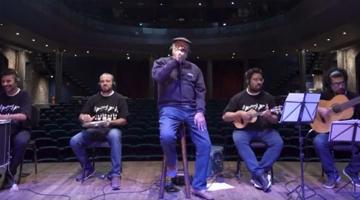 quatro músicos tocando instrumentos sentados e um ao centro cantando. #paratodosverem