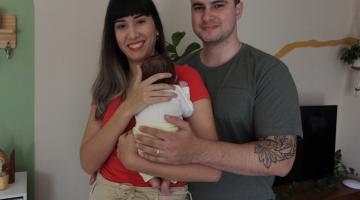 casal posa para foto. A mulher segura o bebê. Com parte do corpo atrás da mulher, homem coloca a mão sobre o corpinho do bebê. #paratodosverem