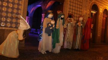 Atores vestidos de bonecos encenam Maria com o menino Jesus no colo, José, um anjo, e dois reis magos ao fundo. #paratodosverem