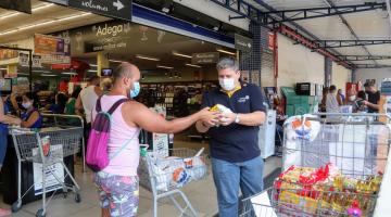 Homem de camisa branca, com carrinho de supermercado, entrega doação para homem de camisa azul em frente a supermercado. #pracegover