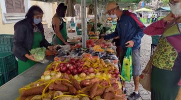 banca em feira com pacotes de batatas doces, cebolas e outros itens. Pessoas circulam em torno.  #paratodosverem