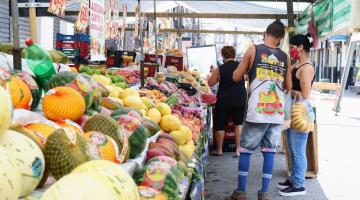 barraca de feira com frutas ao lado esquerdo. Feirante e consumidora do lado direito. Ele está de costas e ela, de lado, segurando uma sacola com bananas. #paratodosverem