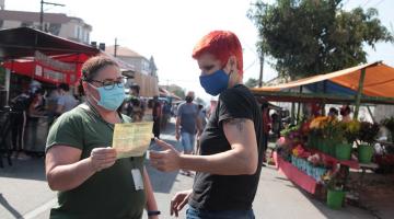 mulher entrega folheto a outra mulher no meio da feira. #paratodosverem