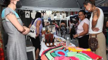 Mulheres observam produtos de crochê em estande. #pratodosverem