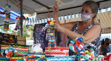 Mulher segura boneco feito de tampas de garrafa. Várias objetos com a mesma peça estão expostos em banca. #Paratodosverem