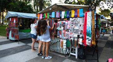 Três mulheres manuseiam artigos em barraca. #Paratodosverem