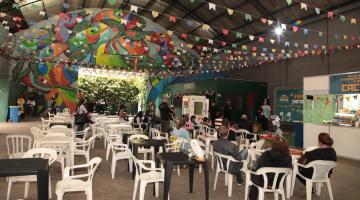 galpão decorado com bandeirinhas, mesas e cadeiras dispostas com algumas pessoas sentada em torno