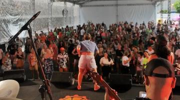 cantor se apresenta em palco com público à frente, Todos estão sob uma grande tenda. #paratodosverem 
