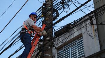 Profissional no alto de escada mexe em fiação em poste. #pracegover