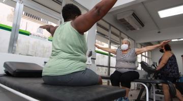 duas mulheres estão sentadas em macas fazendo movimentos com os braços. Eles estão estendidos nas laterais. #paratodosverem