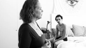 cewna de fim. Uma mulher, de lado, está em pé. Sem olhar diretamente, ela fala com um homem que está sentado em uma cama. #paratodosverem