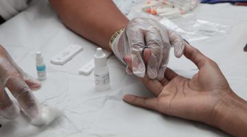 Mãos com luvas seguram a mão de alguém que acaba de fazer o teste rápido. A ponta do dedo do examinado tem uma gota de sangue. Há frascos pequenos na mesa. #Paratodosverem