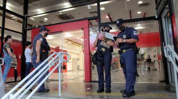 Agentes estão na porta de entrada de um banco. Eles têm bloco de anotações nas mãos. #Paratodosverem