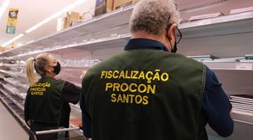 dois fiscais do procon uniformizados são fotografados de costas. Eles estão diante de prateleiras em papelaria observando produtos. #paratodosverem