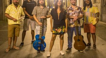 os seis integrantes da banda, alguns segurando instrumentos como sino, guitarra, violão,  pandeiro. A mulher está à frente. #paratodosverem