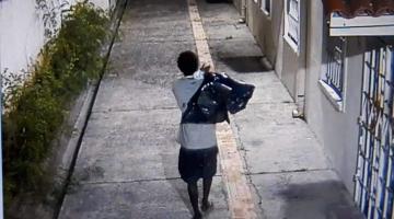 Imagem captada via câmera de monitoramento mostra homem andando em calçada, de costas para a imagem e carregando dois sacos plásticos nos ombros. #Paratodosverem