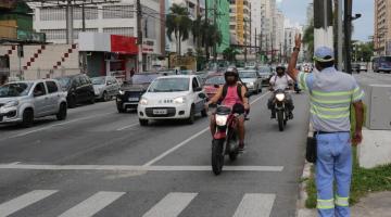 Agente da CET está no canteiro central de avenida sinalizando para motoristas. Ele está ao lado de uma faixa de pedestres. #Paratodosverem