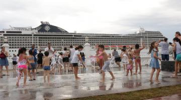 Crianças brincam como água da fonte interativa com navio de cruzeiro passando ao fundo. #pracegover