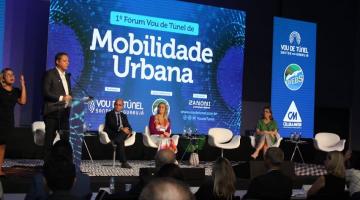 palco com pessoas sentadas e o prefeito em pé falando ao público. Ao fundo, um painel onde se lê Mobilidade Urbana - #paratodosverem