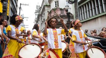 Grupo vestido com roupas do tipo afro e instrumentos de percussão desfila em rua. #Pracegover