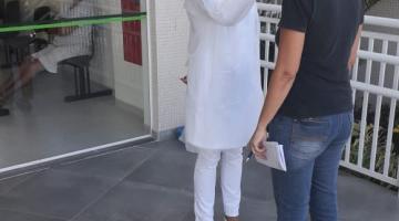 Mulher vestida de branco, usando luva, touca e máscara mede temperatura de uma mulher. Ambas estão em pé na porta de entrada de um prédio. A mulher de branco aponta um termômetro para a testa da outra. #Paratodosverem