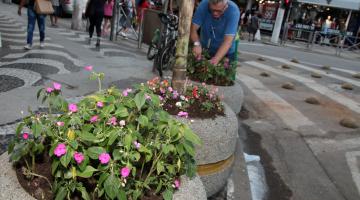 Vasos com flores em praça. Ao fundo, um funcionários planta mudas em outro vaso. #Pracegover