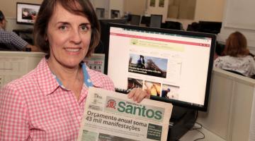 Márcia está segurando uma antiga edição do Diário Oficial em papel. Atrás dela, um monitor de desktop está aberto no  Santos Portal. #Pracegover