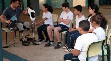 Pauê está sentado em uma cadeira usando e mostra exemplar de próteses para os alunos. Uma das meninas, ao lado dele, toca na peça. Os outros estudantes observam. #Pracegover