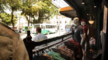 Imagem feita de dentro de loja com vista para uma praça. Dentro da loja estão camisas penduradas em araras e manequins com roupas. Na rua passa uma pessoa e um ônibus ao fundo. #Pracegover