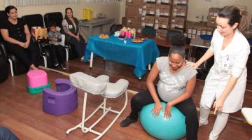 Mulheres estão reunidas em uma sala onde estão expostos diversos produtos para o conforto na hora do parto. À direita, uma gestante está sentada em uma bola de pilates. #Pracegover