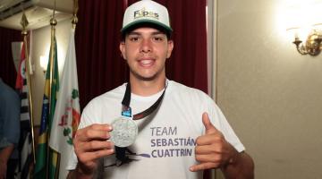 o canoísta segura com a mão direita a medalha que está pendurada no pescoço. #pratodosverem