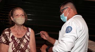 Mulher recebe vacina no braço esquerdo aplicada por uma pessoa que está de branco #paratodosverem