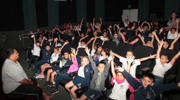 crianças estão em sala de cinema felizes, todas com os braços para o alto. #paratodosverem 