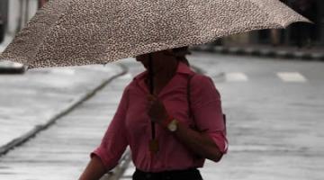 mulher atravessa rua sobre trilhos do bonde segurando guarda-chuva. Está chovendo. #paratodosverem 