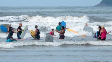 pessoas maduras entram no mar com pranchas. #paratodosverem 