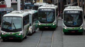 Seis ônibus em uma rua. Eles fazem fila à esquerda e outros dois estão passando ao lado. #Pracegover 