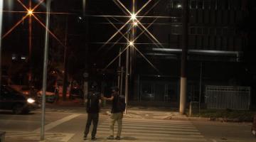 duas pessoas estão atravessando faixa iluminada à noite. #paratodosverem 