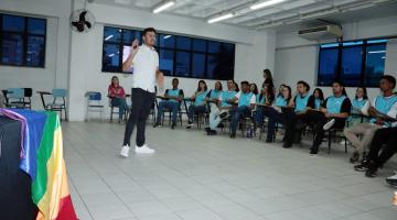 em primeiro plano, mesa com bandeira da causa lgbt+. Ao fundo, homem em pé da palestra para jovens sentados. #paratodosverem 