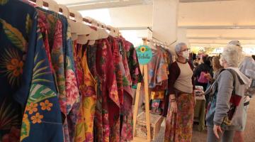 roupas coloridas penduradas em arara em ambiente de feira criativa. #paratodosverem 