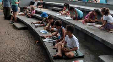 crianças estão sentadas pintando figuras desanhadas em papel. #paratodosverem